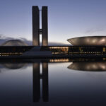 Fachada do Congresso Nacional ao entardecer, com as duas torres, a cúpula da Câmara dos Deputados e o prato invertido do plenário do Senado refletidos no espelho d’água