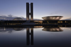 Fachada do Congresso Nacional ao entardecer, com as duas torres, a cúpula da Câmara dos Deputados e o prato invertido do plenário do Senado refletidos no espelho d’água