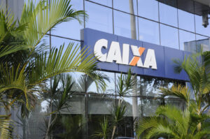 Fachada de vidro de agência da Caixa Econômica Federal com o logotipo azul e laranja da Caixa