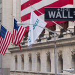 Placa da Wall Street em frente ao edifício da Bolsa de Nova York, com bandeiras dos Estados Unidos